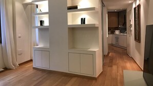 A modern living room with built-in shelving, a flat-screen TV, and a kitchen in the background. - SAN DOMENICO LUXURY APARTMENT (Siena)