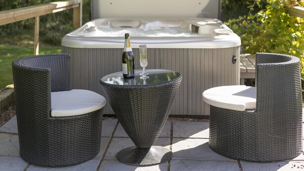 A hot tub with a wooden deck, two wicker chairs, and a table with a bottle and glass. - The Pines at Eastleigh - 3 Cottages (Bideford)