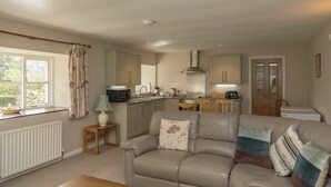 A cozy living room with a sofa, a small table, and a kitchen in the background. - The Pines at Eastleigh - 3 Cottages (Bideford)