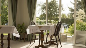 A dining area with a table and chairs, a view of greenery outside, and a large window. - The Pines at Eastleigh - 3 Cottages (Bideford)