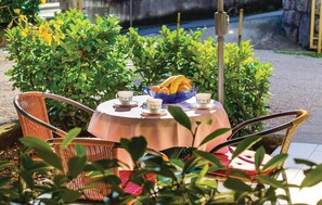 Uma mesa redonda com toalha branca, duas cadeiras de vime, uma tigela azul com frutas e chávenas, e vegetação ao fundo.