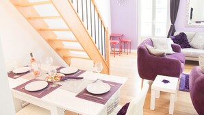 A dining area with a table set for four, a staircase, a purple sofa, and a small white table. - SunlightProperties LEGEND: 2 Bedroom Rental, near Beach with balcony & AC (Nice)