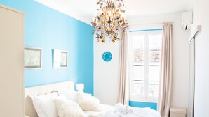 A bedroom with a white bed, blue walls, a chandelier, a clock, and a window with curtains. - SunlightProperties LEGEND: 2 Bedroom Rental, near Beach with balcony & AC (Nice)
