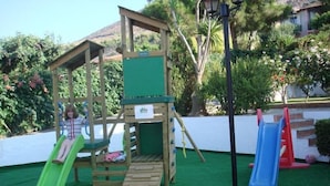A wooden play structure with a green slide and a blue slide, surrounded by greenery and a white wall. - Sunrise Apartments Hersonissos (Hersonissos)