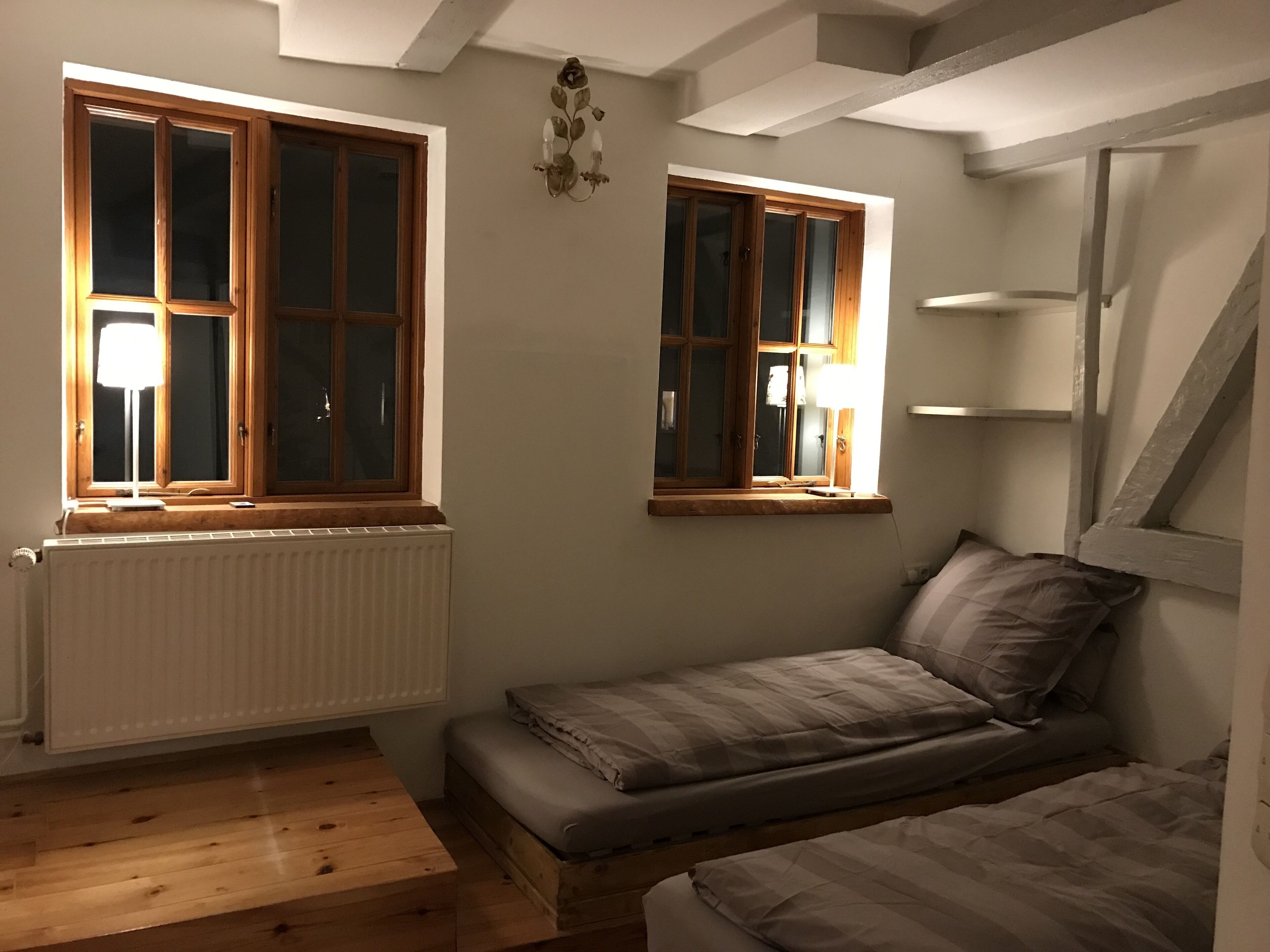 Una stanza piccola e accogliente con due finestre, un termosifone, un pavimento in legno e un letto.
