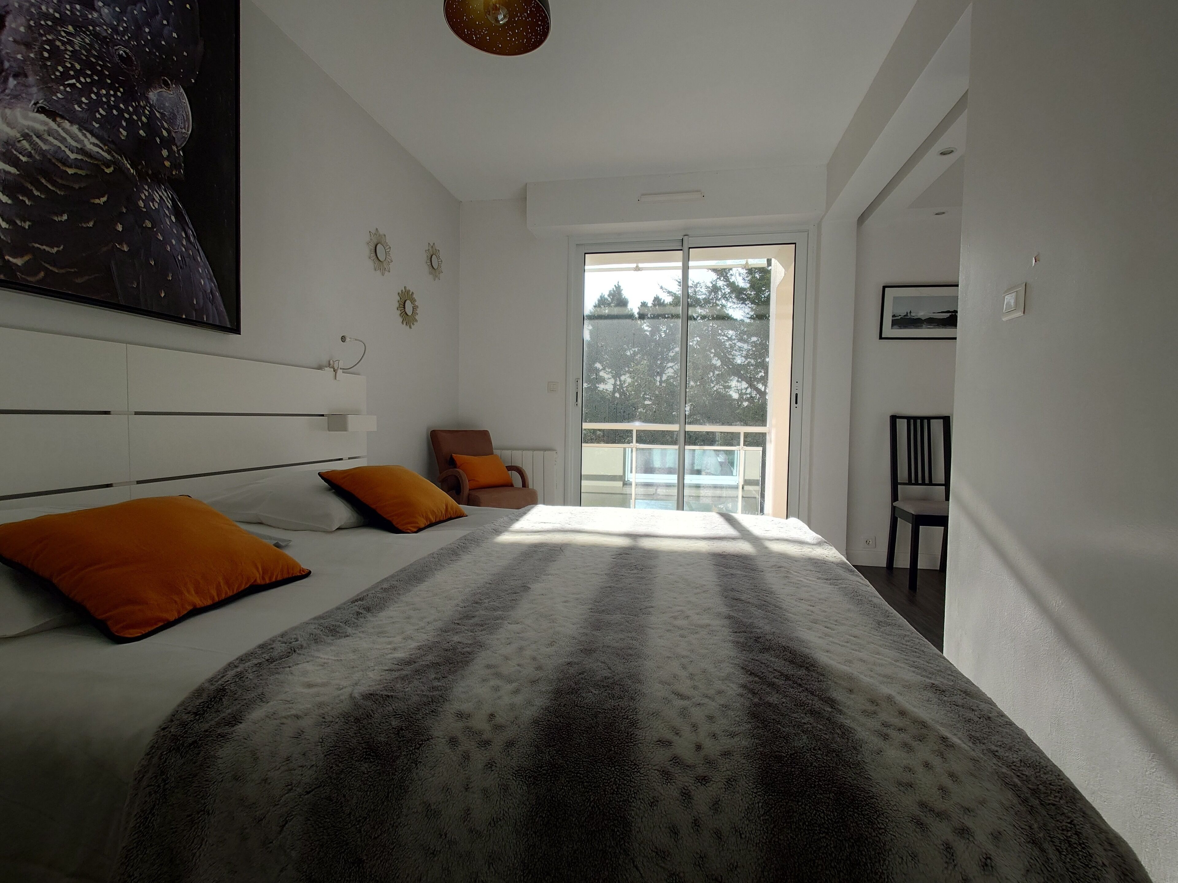Un dormitorio moderno con una cama grande, dos almohadas naranjas, vistas al exterior a través de una puerta corrediza y una lámpara en el techo.