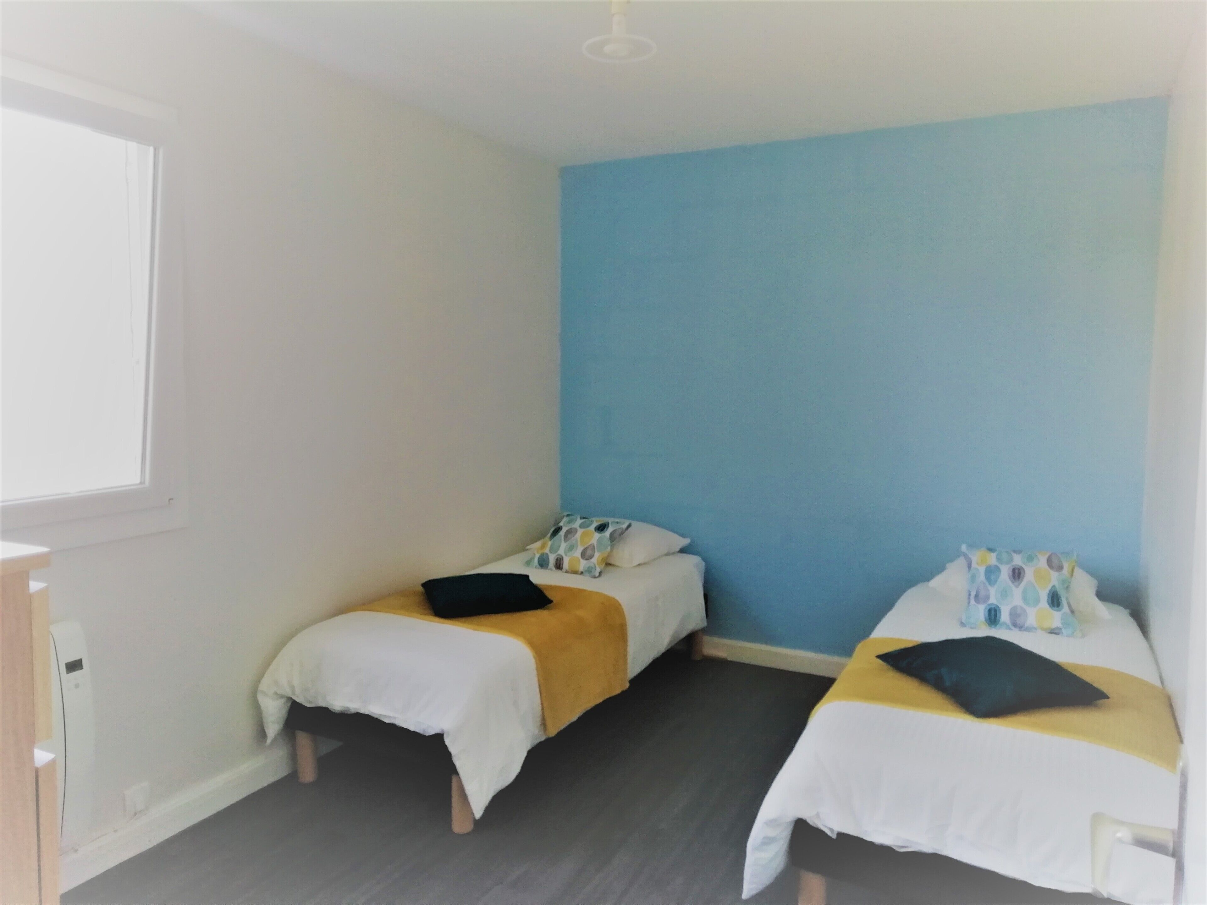 Una habitación pequeña con dos camas individuales, cada una con un edredón blanco y una almohada. La habitación tiene una pared azul decorativa y una ventana a la izquierda.