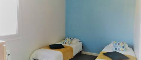 Una habitación pequeña con dos camas individuales, cada una con un edredón blanco y una almohada. La habitación tiene una pared azul decorativa y una ventana a la izquierda.