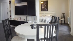 Une chambre d’hôtel moderne avec une table de salle à manger ronde, des chaises noires, un lit avec une tête de lit blanche, un téléviseur fixé au mur et une œuvre d’art encadrée accrochée au mur.