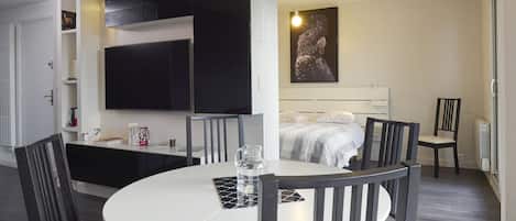 A modern hotel room with a round dining table, black chairs, a bed with a white headboard, a mounted TV, and a framed artwork on the wall.