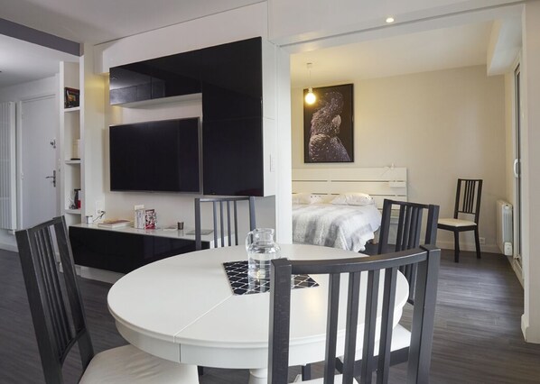 Une chambre d’hôtel moderne avec une table de salle à manger ronde, des chaises noires, un lit avec une tête de lit blanche, un téléviseur fixé au mur et une œuvre d’art encadrée accrochée au mur.