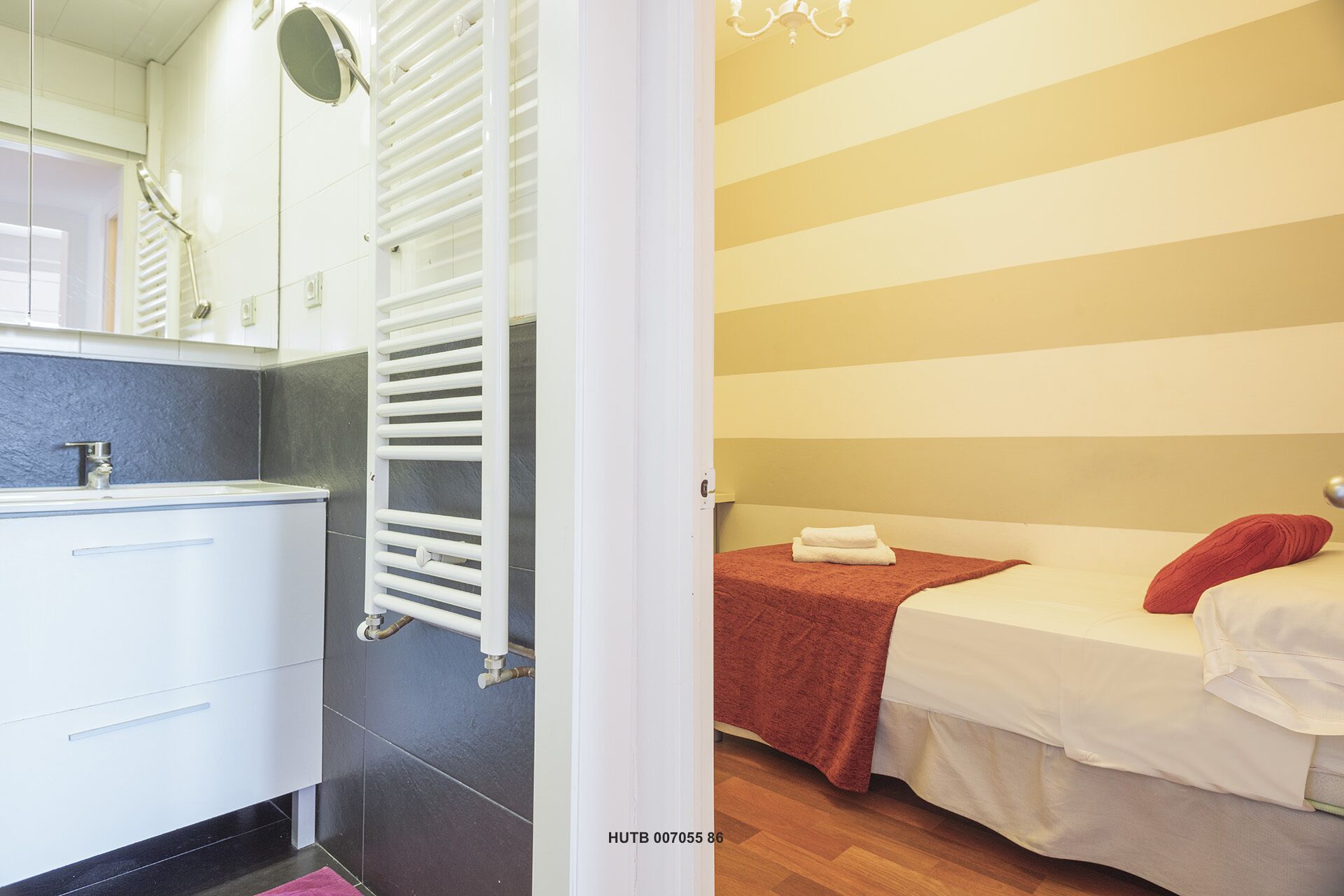 A hotel room with a bathroom featuring a sink, mirror, and towel rack. The room has a bed with a red blanket and pillows, and a wooden floor.