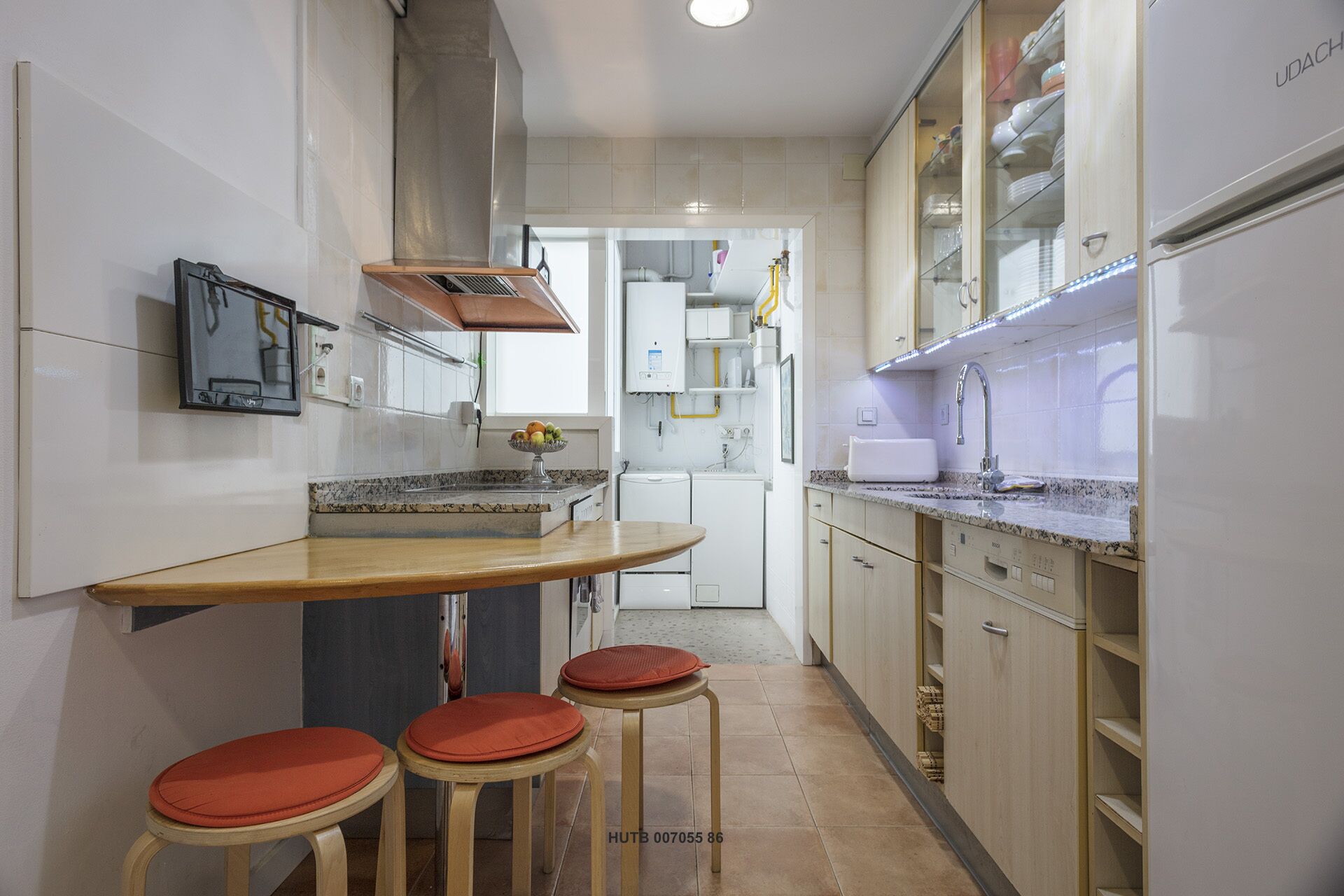 Une cuisine étroite équipée d’un four encastré, d’un évier et d’un plan de travail. On y trouve des tabourets et une petite table sur laquelle est posé un panier de fruits.
