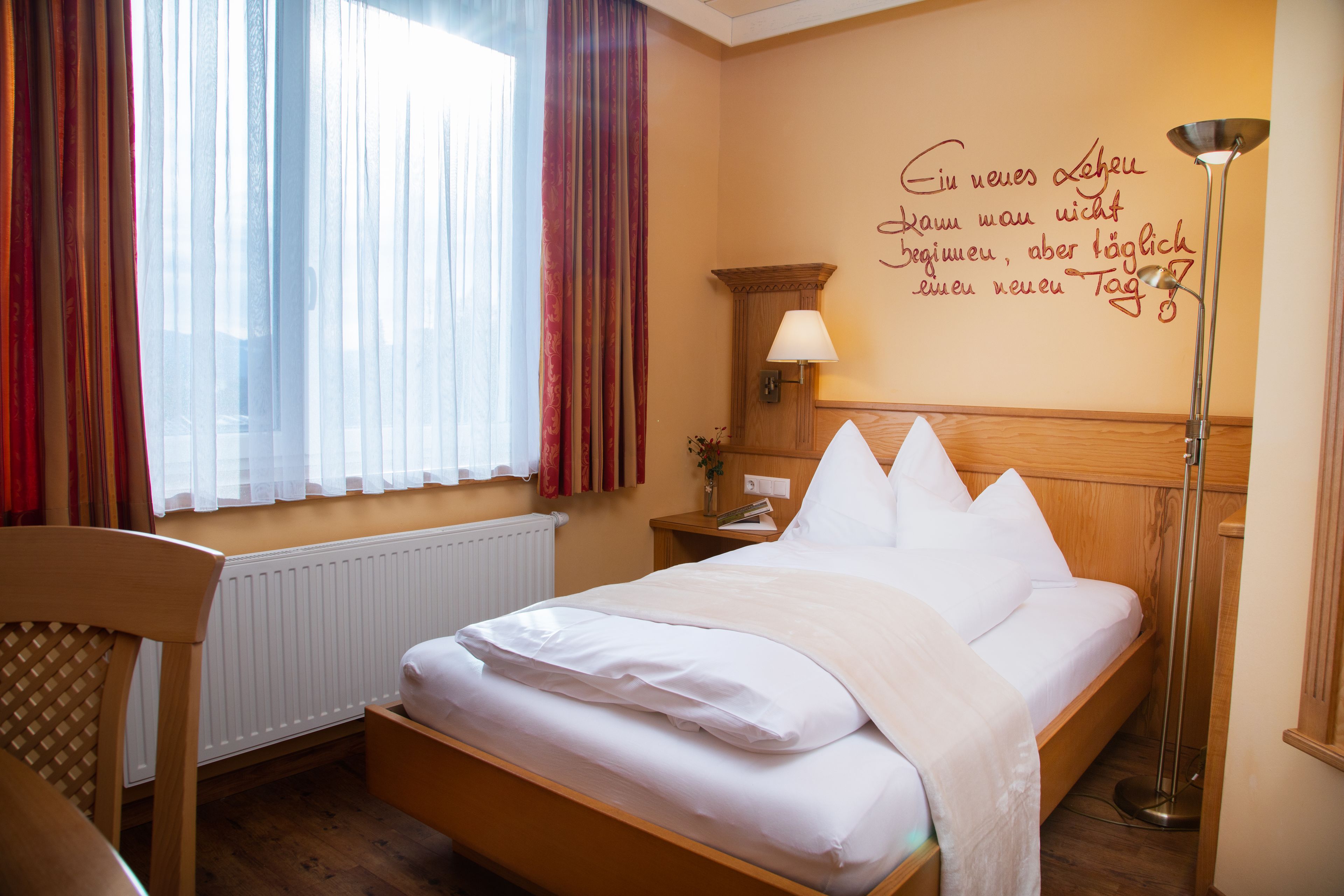 A neatly made bed with white linens in a room with a wooden headboard, a bedside table with a lamp, and a window with sheer curtains.
