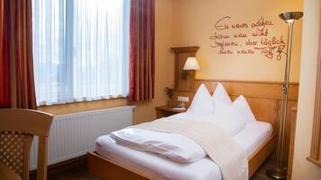 A neatly made bed with white linens in a room with a wooden headboard, a bedside table with a lamp, and a window with sheer curtains.