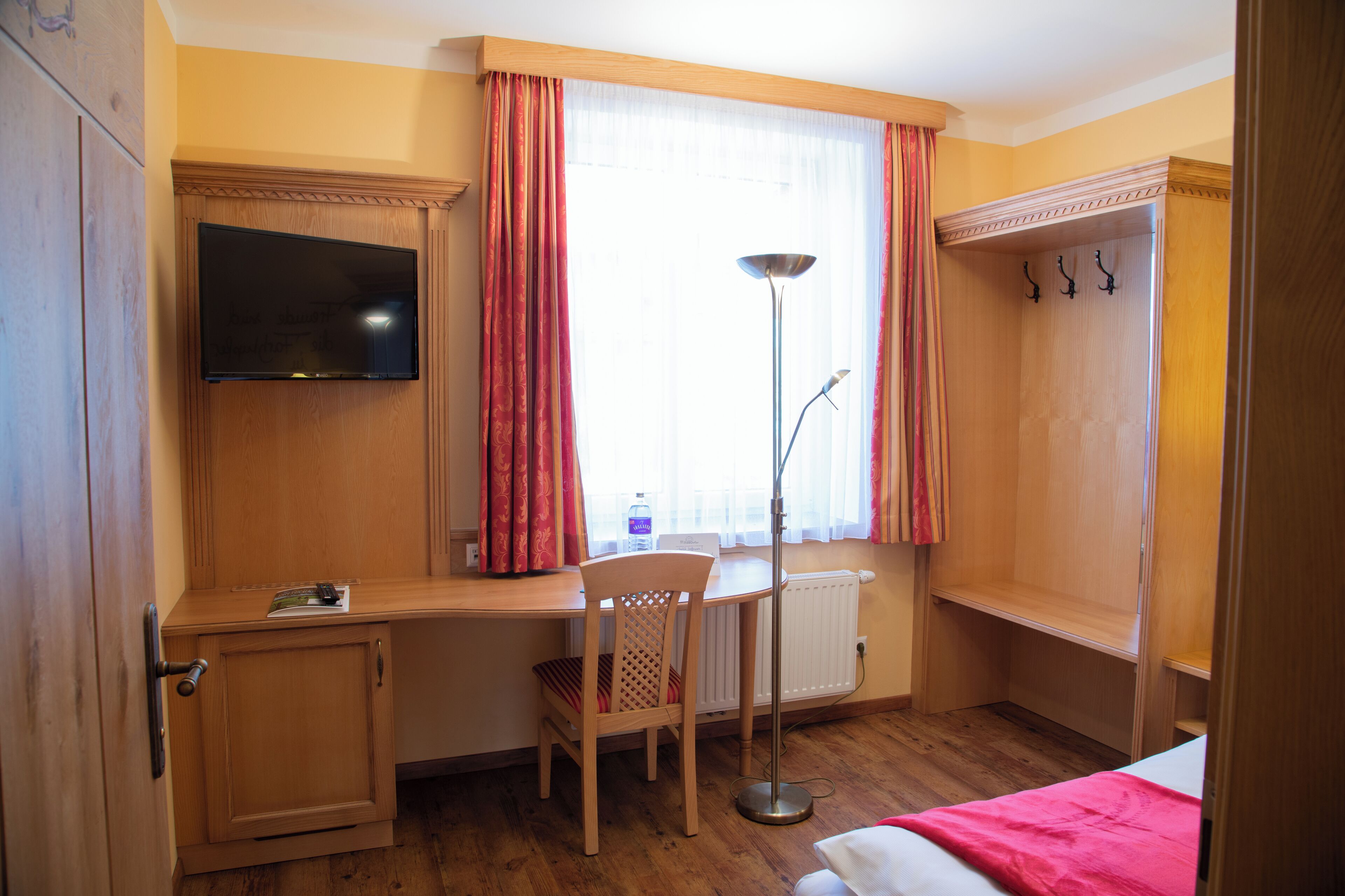 Een hotelkamer met een houten bureau, een stoel, een bed, een aan de muur bevestigde televisie, een vloerlamp en een radiator.