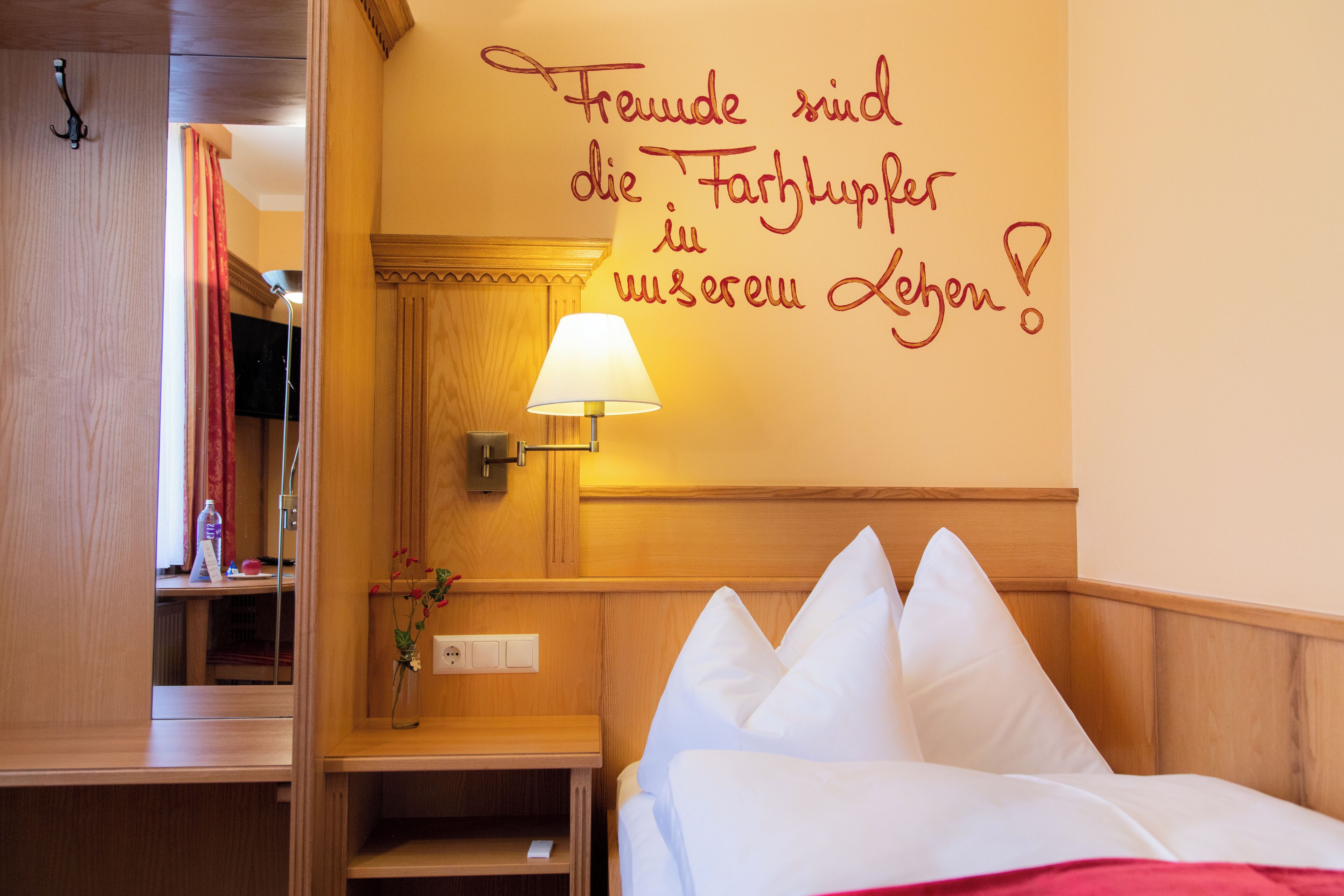 A hotel room with a bed, a nightstand, a lamp, a mirror, and a wall with a German phrase.