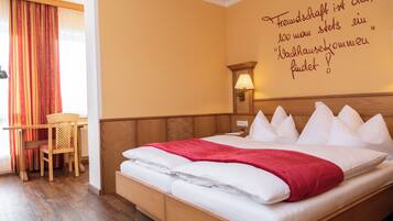 A hotel room with a wooden bed, white pillows, a red blanket, a wooden desk, a chair, and a wall with a German quote.