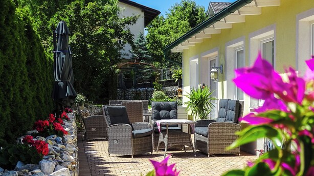 An outdoor patio with wicker furniture, a stone wall, and a yellow building in the background. - Bed & Breakfast Mittelkärnten (Althofen)