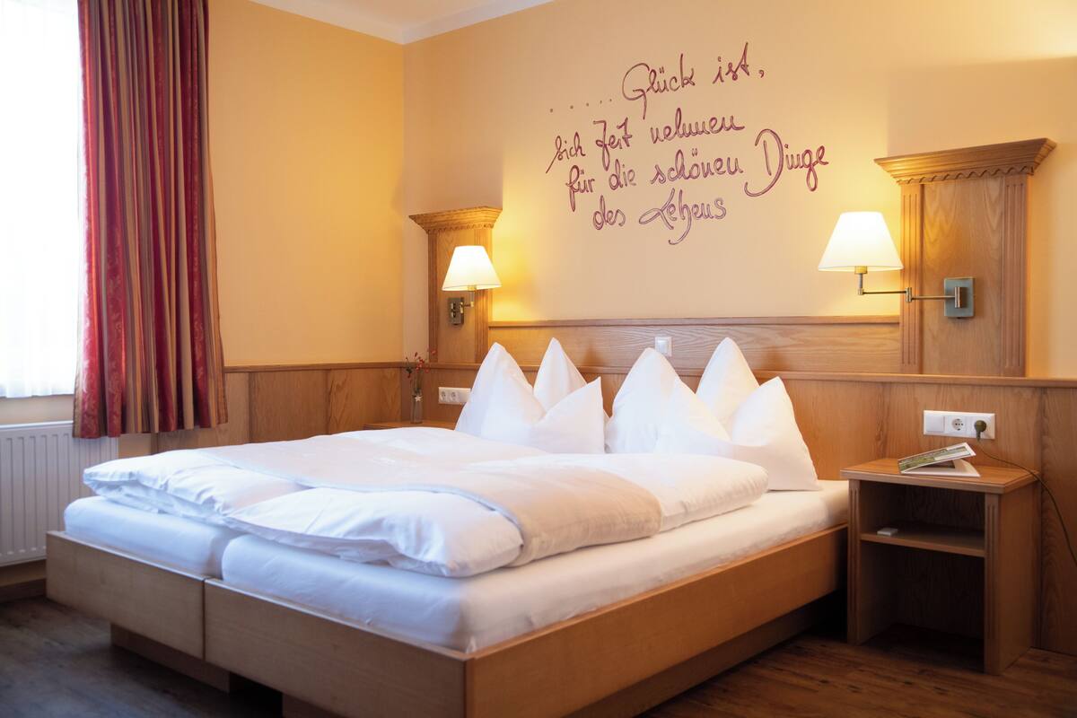 A hotel room with a large bed, two bedside lamps, a wooden headboard, and a wall with a German phrase.