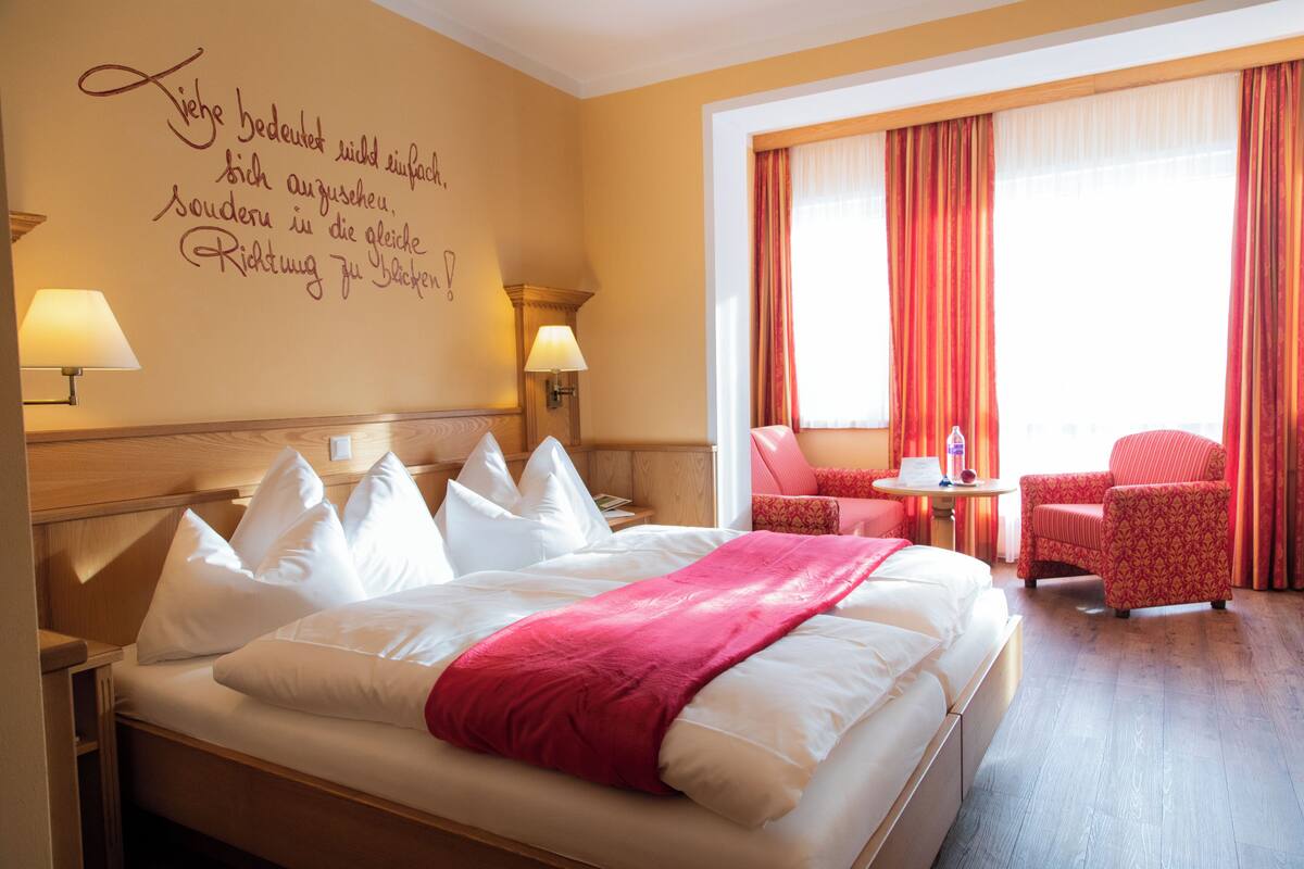 A hotel room with a large bed, two bedside lamps, a red chair, a small table, and a window with curtains.
