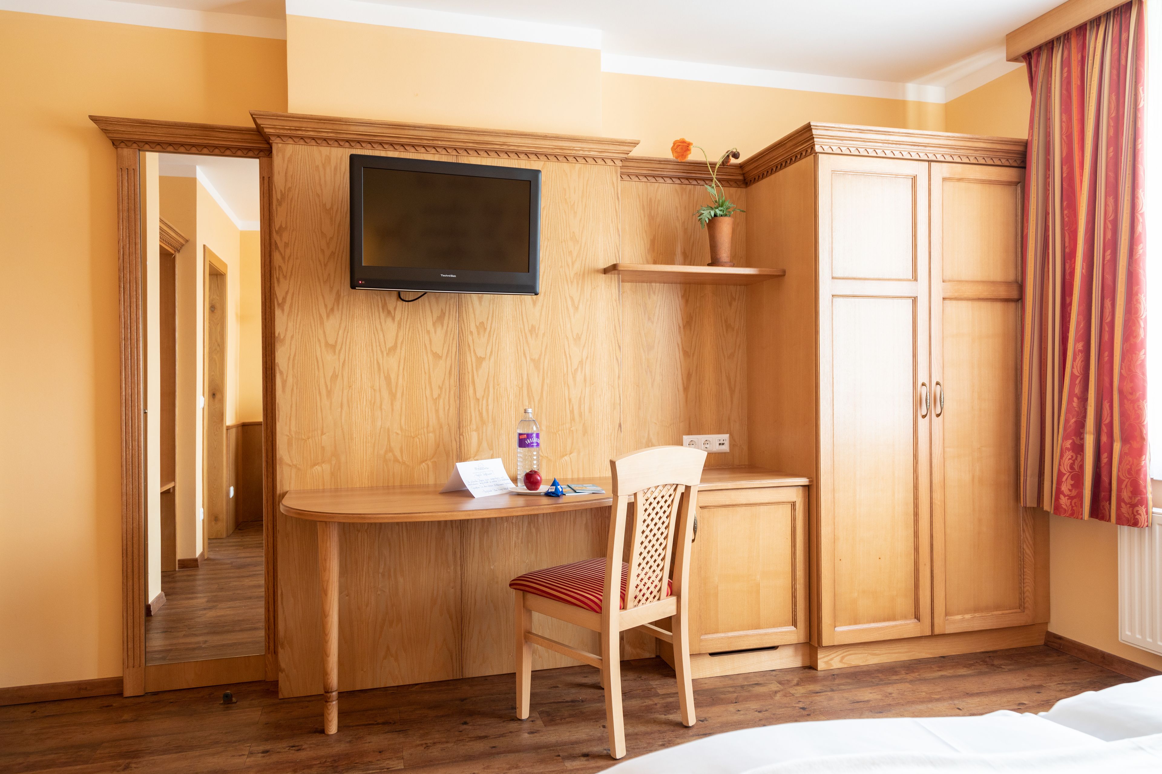 A hotel room with a wooden desk, a chair, a television, a wardrobe, and a mirror.
