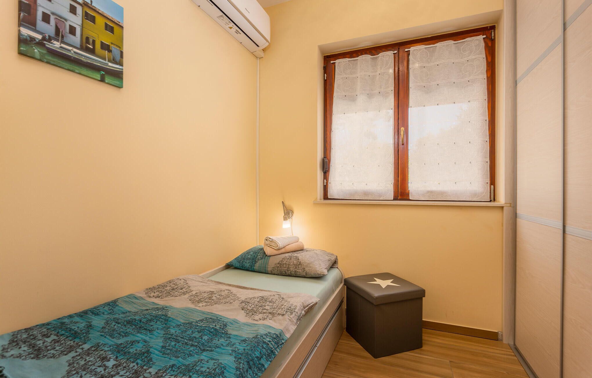 Ein kleines, ordentlich eingerichtetes Schlafzimmer mit Bett, Nachttisch, Lampe und einem Fenster mit Vorhängen.
