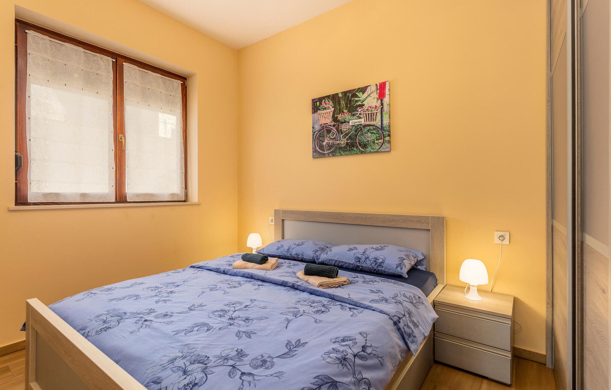 Une chambre à coucher avec un lit, une table de chevet, une fenêtre avec des rideaux et un tableau accroché au mur.