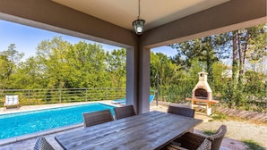 A covered outdoor area with a pool, a wooden table, wicker chairs, and a brick pizza oven. - Gorgeous home in Gostinjac (Gostinjac)