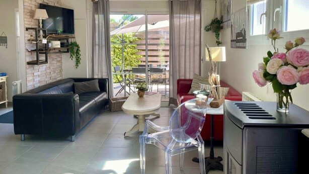 A modern living room with a black leather sofa, a dining table with chairs, a TV mounted on a brick wall, and a kitchen area with a refrigerator. - comfortable house in Lacanau Ocean, everything in walking distance (Lacanau)