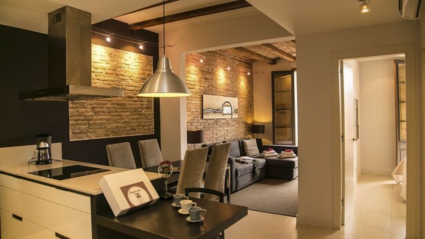 A modern kitchen with a stone wall backsplash, a dining area with a table set for two, and a living room with a sofa and a fireplace. - Alcam Molino (Barcelona)