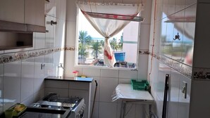 A kitchen with white tiled walls, a gas stove, a window with a view, and a chair. - BUILDING FACING THE SEA IN PRAIA GRANDE-SP (Guilhermina)