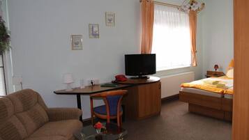 A room with a bed, a desk with a television, a chair, a sofa, a small table with a vase, and a window with curtains.