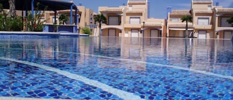 A swimming pool with blue mosaic tiles, surrounded by buildings with beige facades and flat roofs.