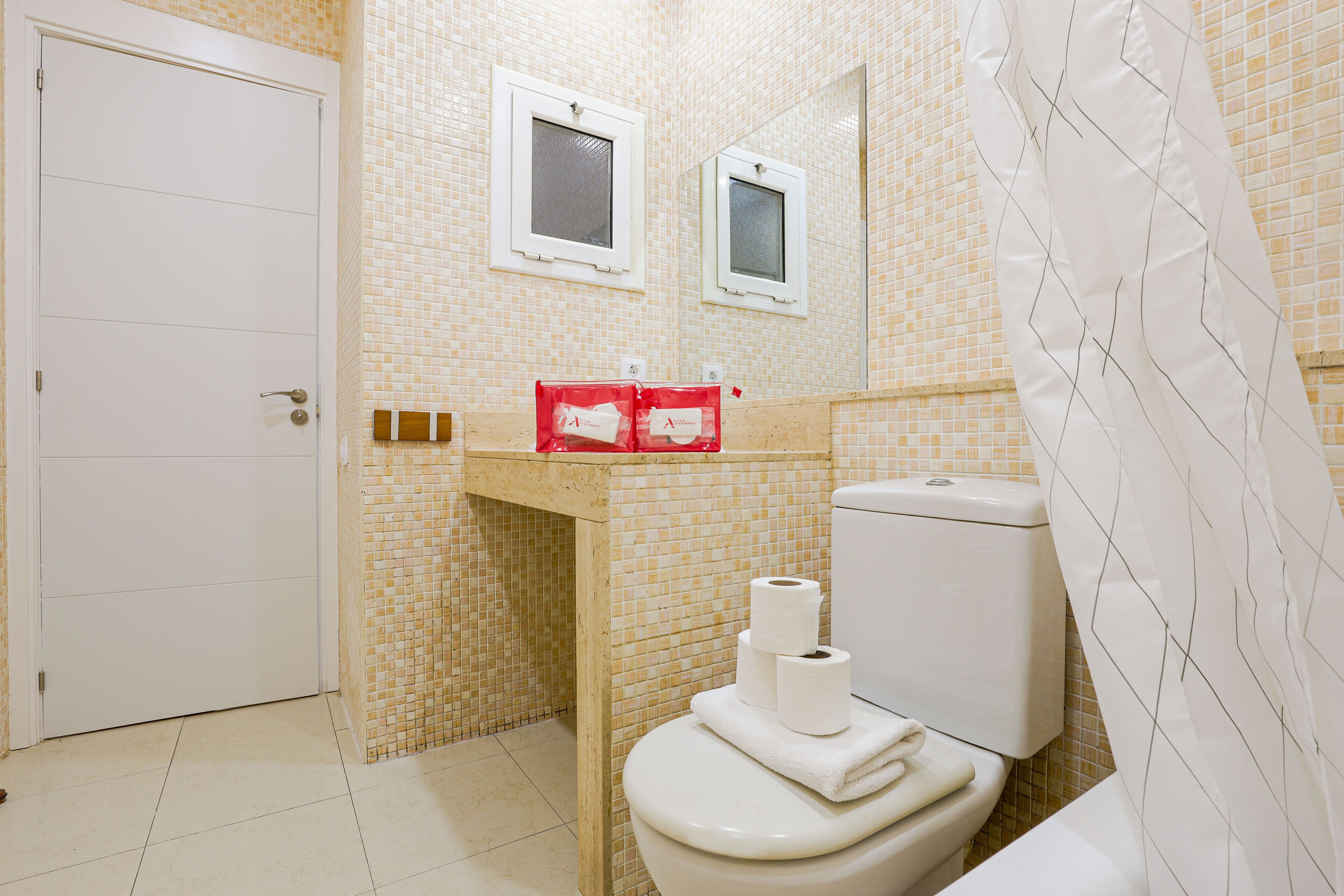 A bathroom with a toilet, a sink, and a mirror. There are two windows on the wall and a shower curtain.