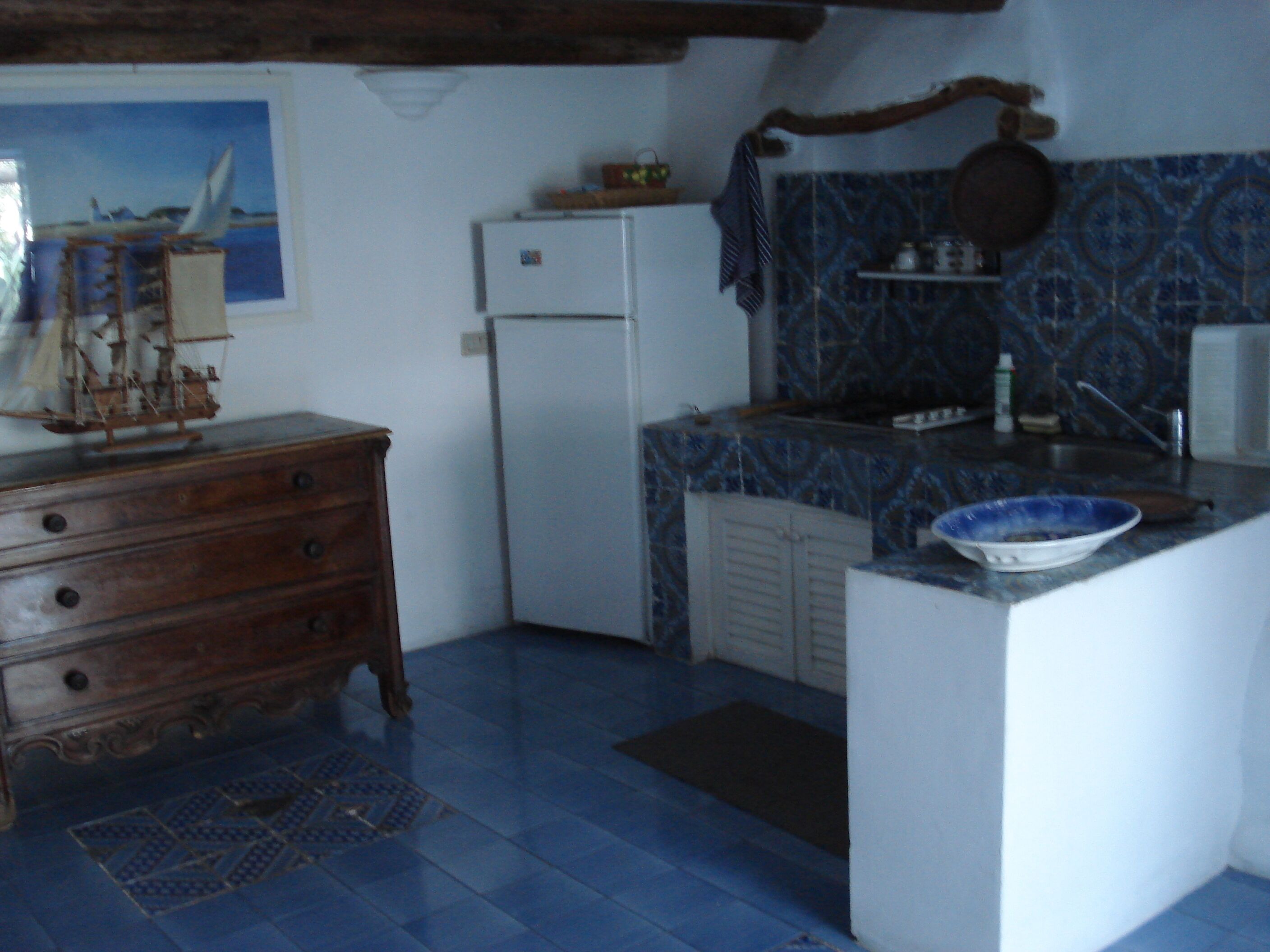 Une cuisine avec un mur recouvert de carreaux bleus, un réfrigérateur blanc, une commode en bois et un petit îlot central sur lequel se trouve un saladier.