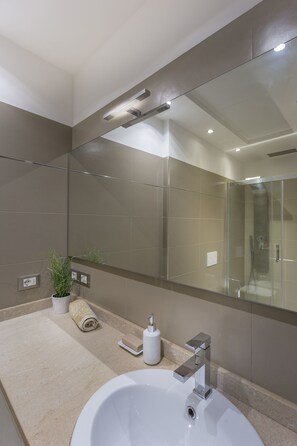 Un baño moderno con un espejo grande, un lavamanos blanco y una ducha con cerramiento de vidrio.
