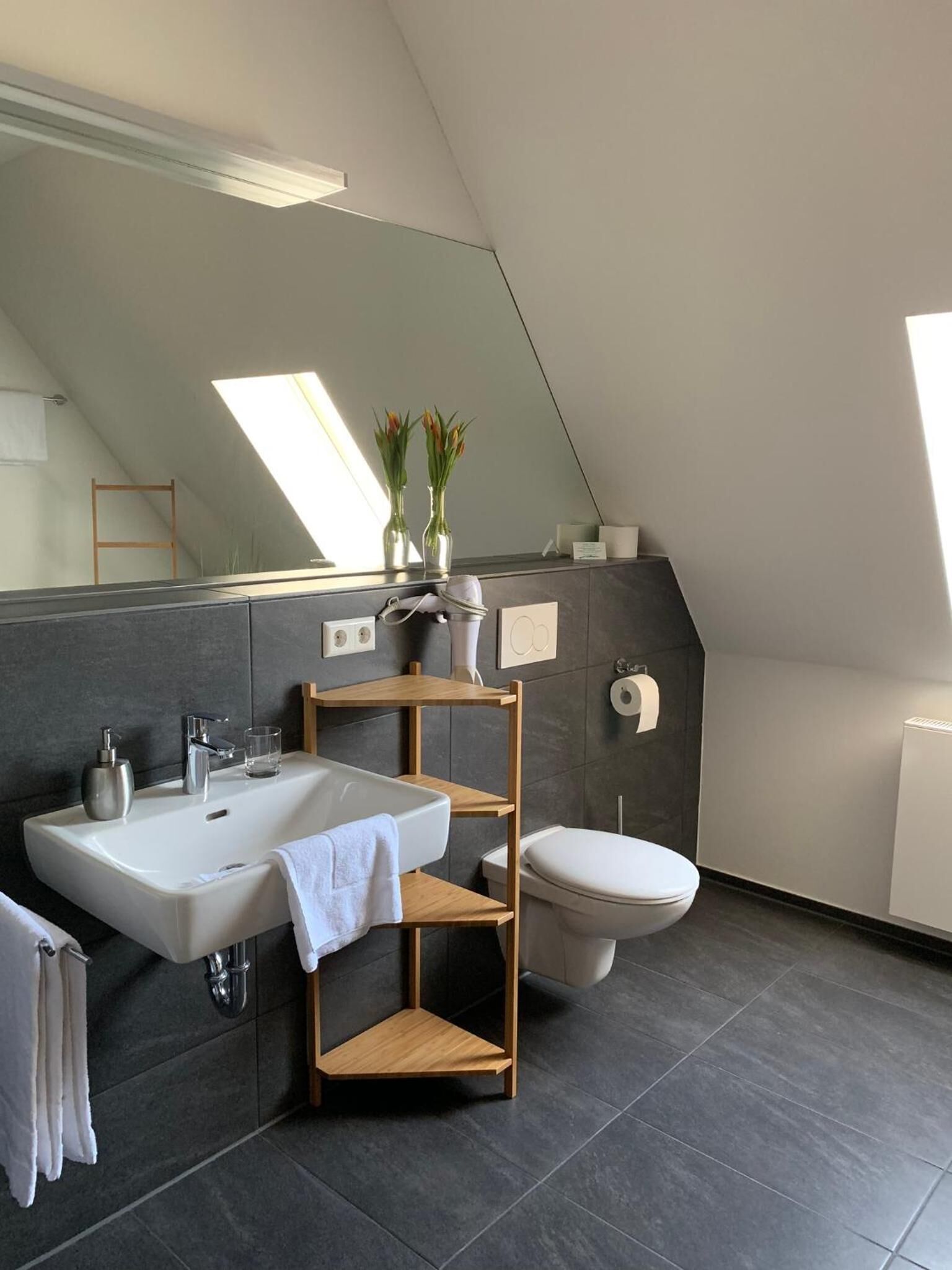 Un baño moderno con un espejo grande, un lavabo, un inodoro y una zona de ducha. Hay un tragaluz sobre el espejo y un perchero con toallas.