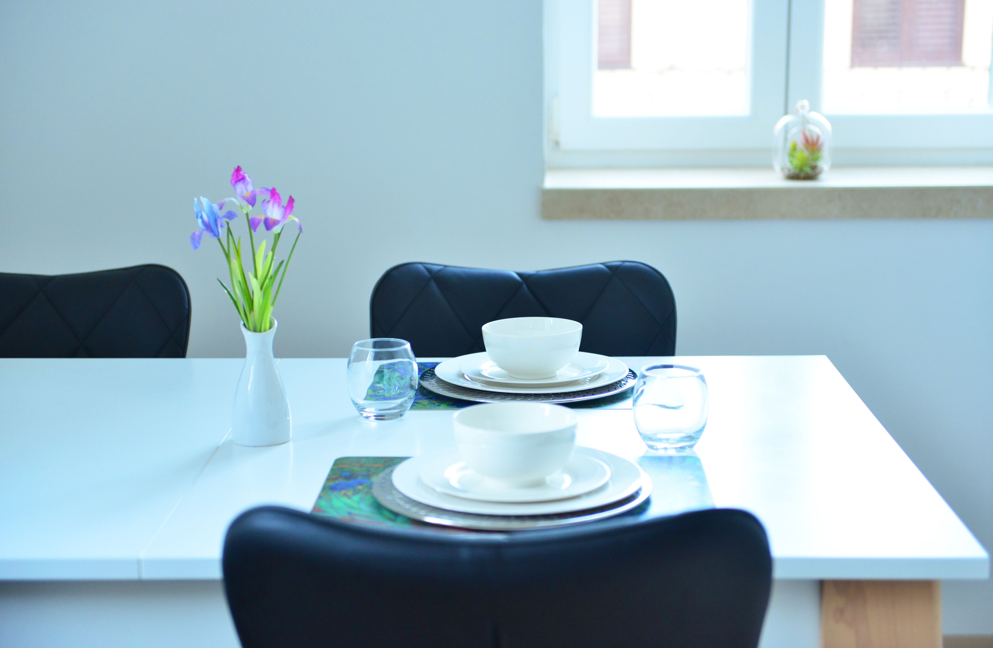 A dining table set with white plates, bowls, and glasses, a vase with flowers, and a window with a view of the outside.