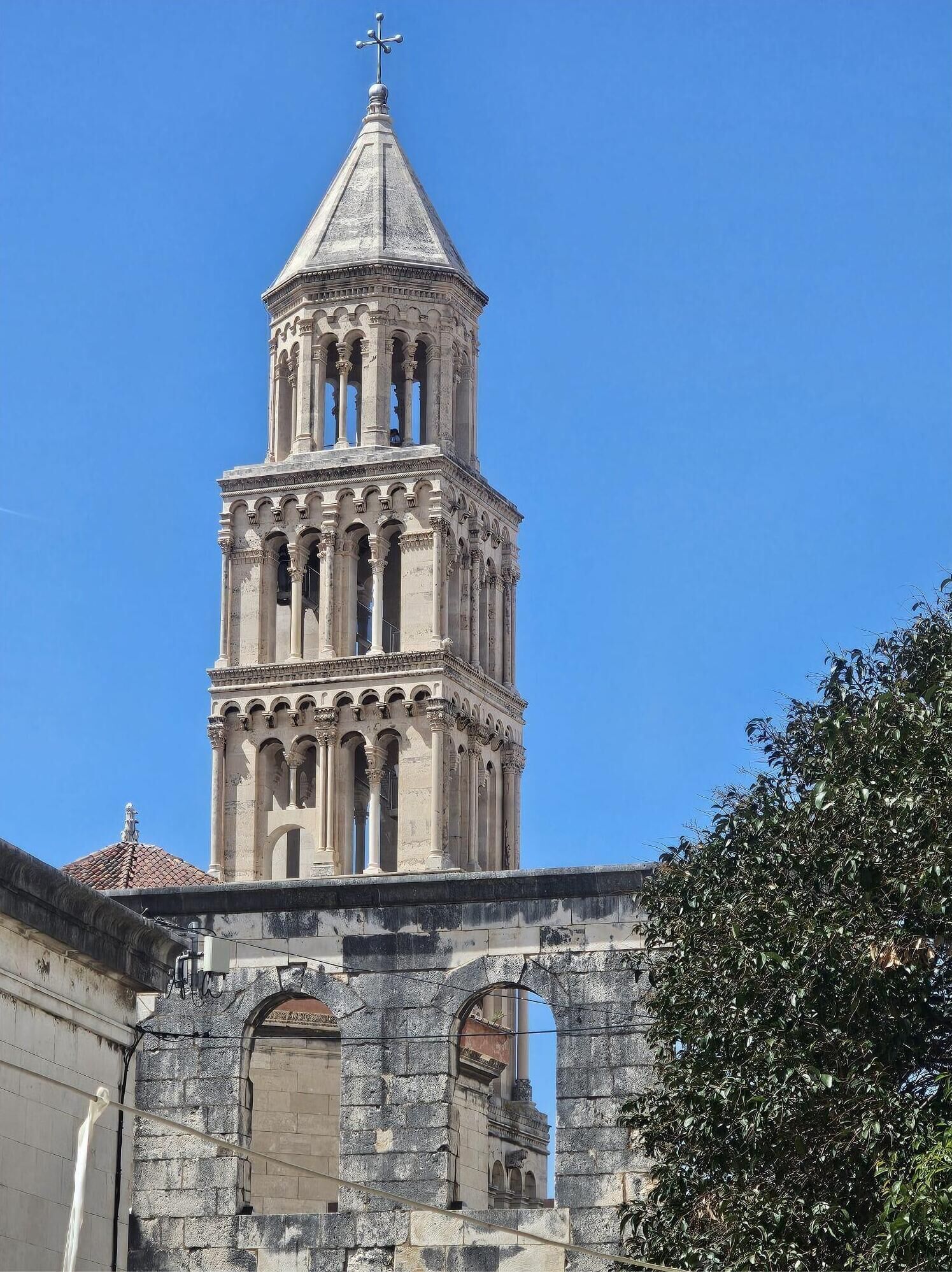 Une tour historique surmontée d’une croix, entourée de murs et d’arcades en pierre.