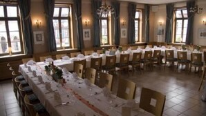 Une salle à manger avec de nombreuses tables dressées pour le repas, de grandes fenêtres avec des rideaux et des luminaires fixés au mur.
