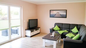 Un salon avec un téléviseur à écran plat posé sur un meuble, un canapé gris avec des coussins verts, une table basse et une grande fenêtre avec des stores.