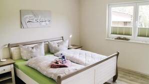 Un lit bien fait, recouvert d’une couvre-lit verte, avec des oreillers blancs et un coussin décoratif orné de motifs floraux. On aperçoit une table de chevet avec une lampe, une œuvre d’art accrochée au mur sur laquelle figure le mot « Relax », et une fenêtre avec des stores.