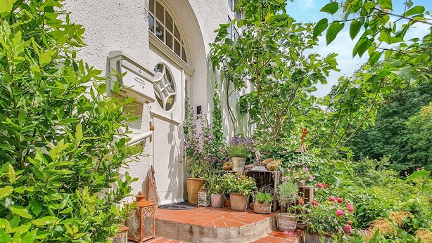 A white building with a large arched window and a door, surrounded by lush greenery and potted plants.