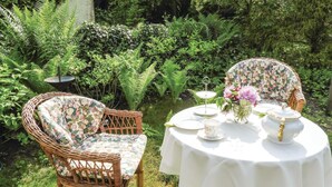 A garden setting with a round table set for tea, surrounded by wicker chairs with floral cushions.