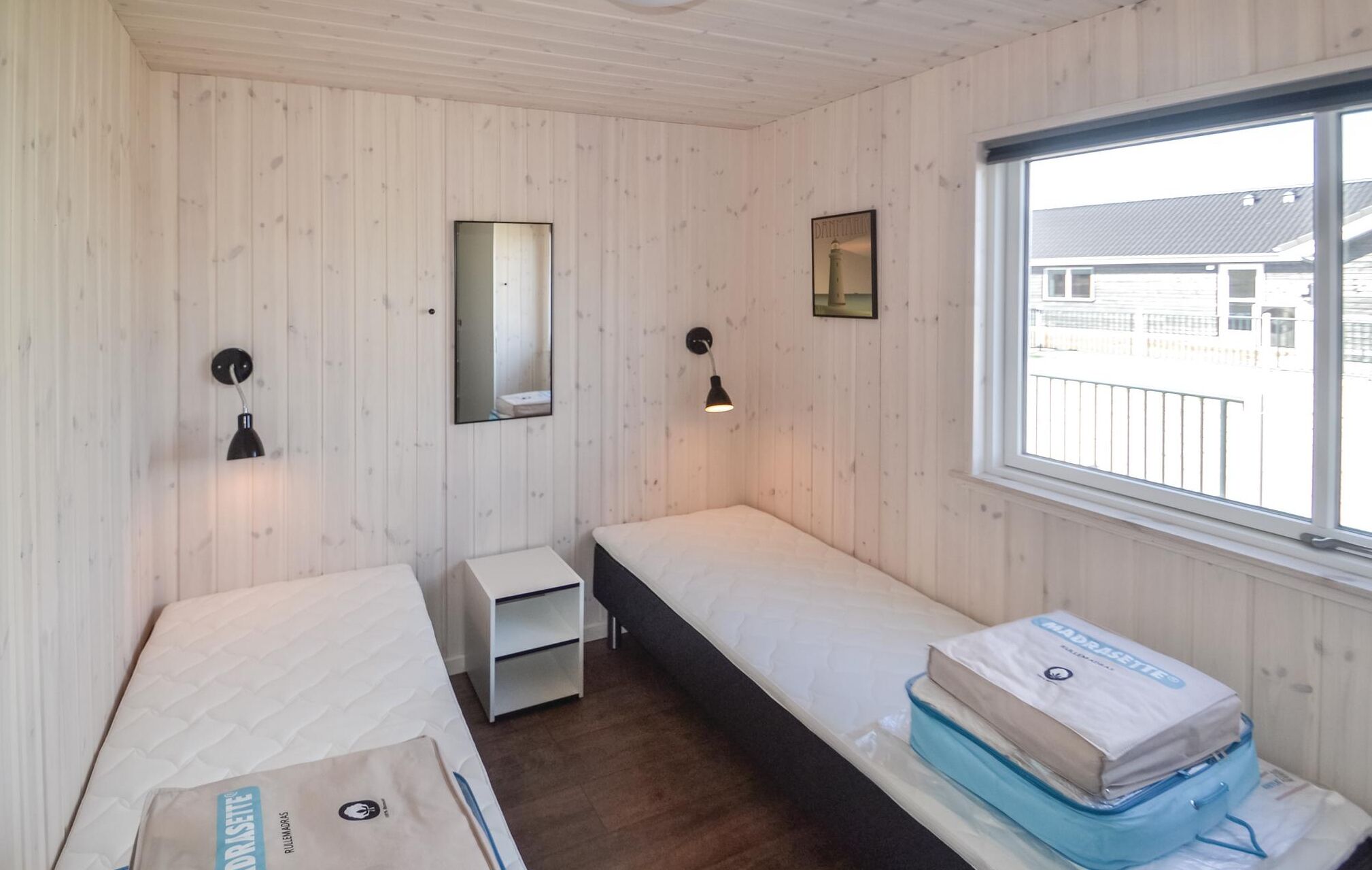 Une petite cabane d’une seule pièce, avec des murs en bois et une fenêtre. On y trouve un lit avec un matelas et un oreiller, une petite table de chevet et un miroir fixé au mur.