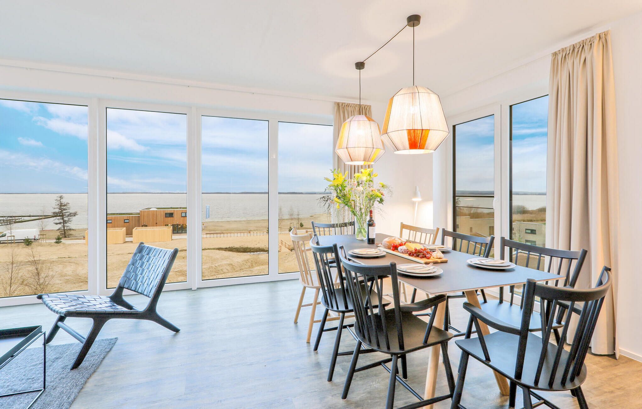 Un comedor con una mesa dispuesta para cuatro, ventanas amplias que ofrecen vistas a la playa y edificios, y dos lámparas colgantes sobre la mesa.