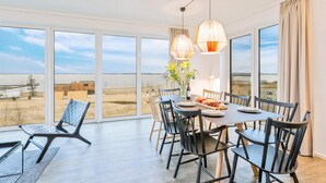 A dining area with a table set for four, large windows offering a view of the beach and buildings, and two pendant lights above the table. - Luxurious apartment in Lembruch/Dümmer See with house sea view (Lembruch/Dümmer See)