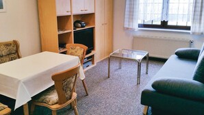 Une pièce avec une armoire en bois, un téléviseur posé sur une étagère, une table recouverte d’une nappe blanche, une table basse en verre et un canapé vert.