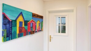 Un couloir avec une porte blanche et un tableau coloré représentant des maisons de plage au mur.